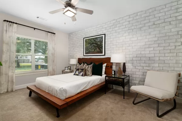 Bedroom with plush carpeting, a queen bed, large window, ceiling fan, end tables, and and arm chair